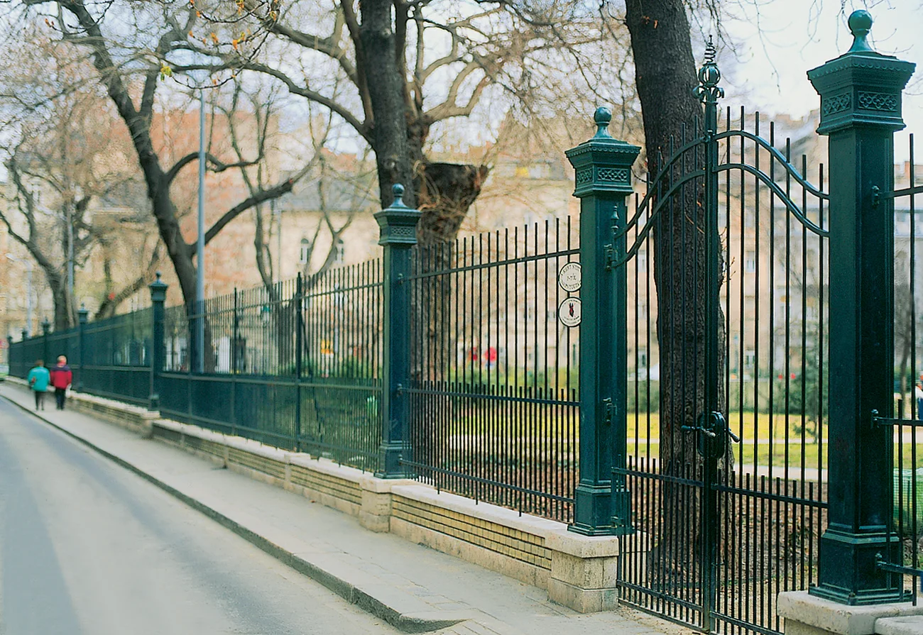 A Károlyi-kert hosszan elnyúló sötétzöld kovácsoltvas kerítése kőlábazaton, szabályos közökben tagolt kerítésoszlopokkal, mögötte a park lombtalan fáival, előtte járda és úttest