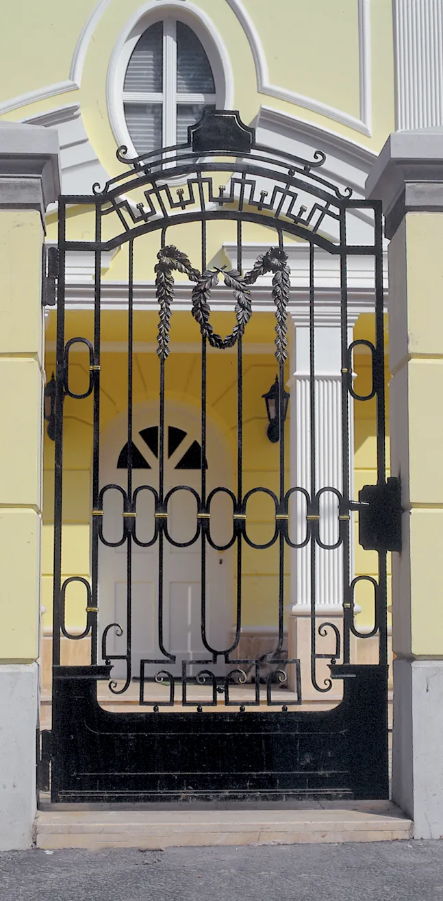 Klasszicizáló épület boltíves kapualjában álló magas kovácsoltvas rácsos kapu, felül meanderes frízzel, alatta girlandmotívummal, a középmezőben ovális gyűrűkből álló kompozícióval