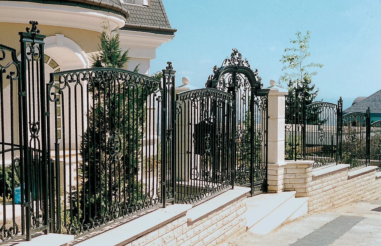 Fekete, díszes kovácsoltvas kerítés és kapu lépcsőzetes kőlábazaton, világos falazott pillérekkel, mögötte oromzatos villa