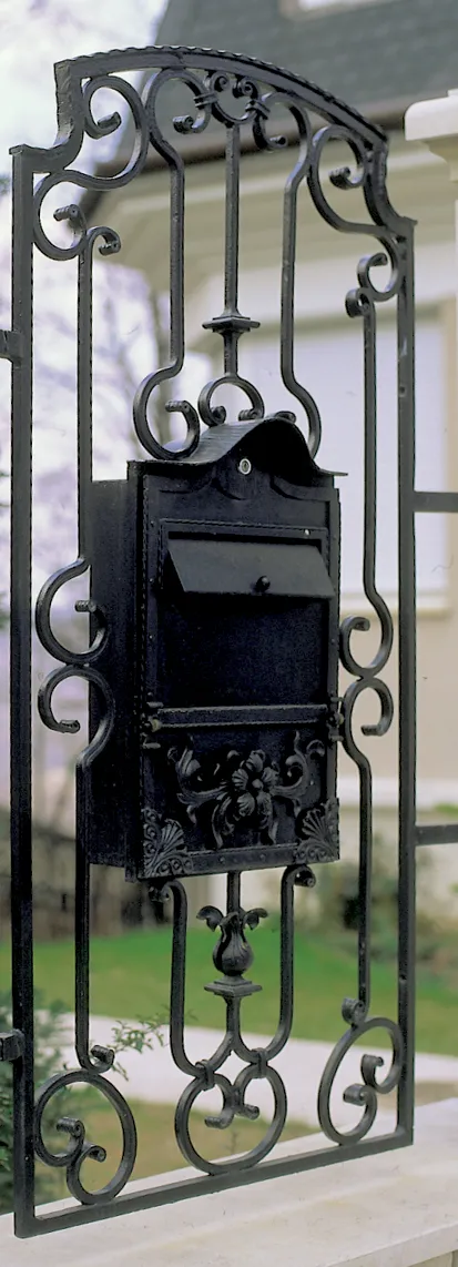 Álló, keskeny kovácsoltvas mezőbe épített fekete postaláda, sátortetős fedéllel, domború levéldíszes előlappal, voluta- és levéldíszes keretben